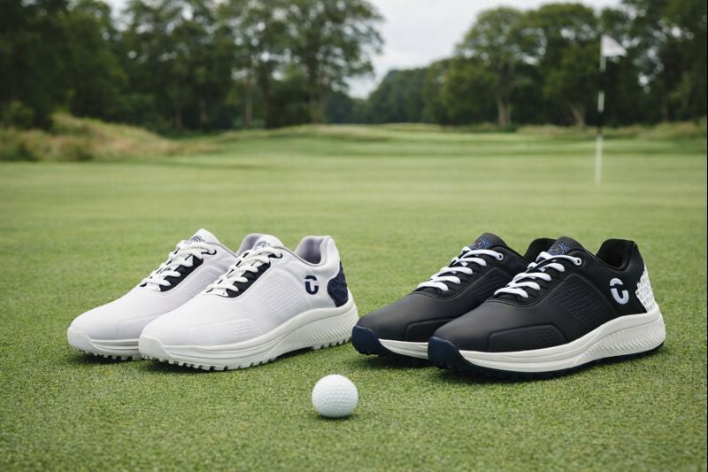 Two pairs of Forza Mens Waterproof golf shoes and a golf ball on a grassy field with trees in the background.